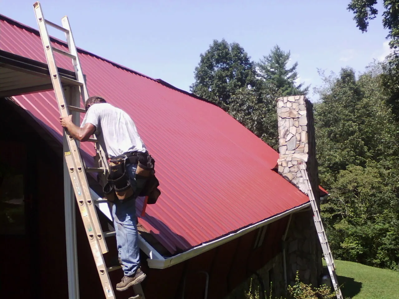 Roofer on a ladder beside a red metal roof and stone chimney — Soffit & Fascia Repair by Roofing Brothers in Bern